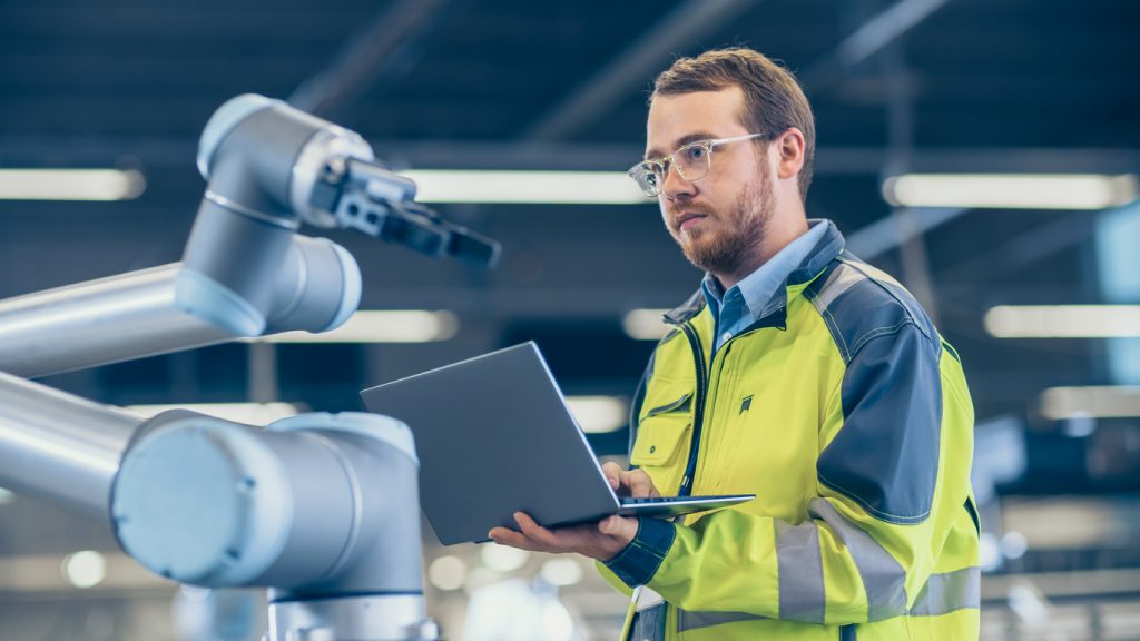 At the Factory: Automation Engineer Uses Laptop for Programming Robotic Arm. New Era in Automatic Manufacturing Industry.